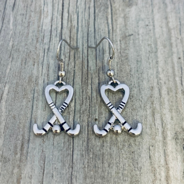 Silver heart-shaped field hockey stick earrings with crossed sticks displayed on rustic wooden surface