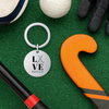 Field hockey LOVE keychain displayed on turf with orange and black sticks and ball