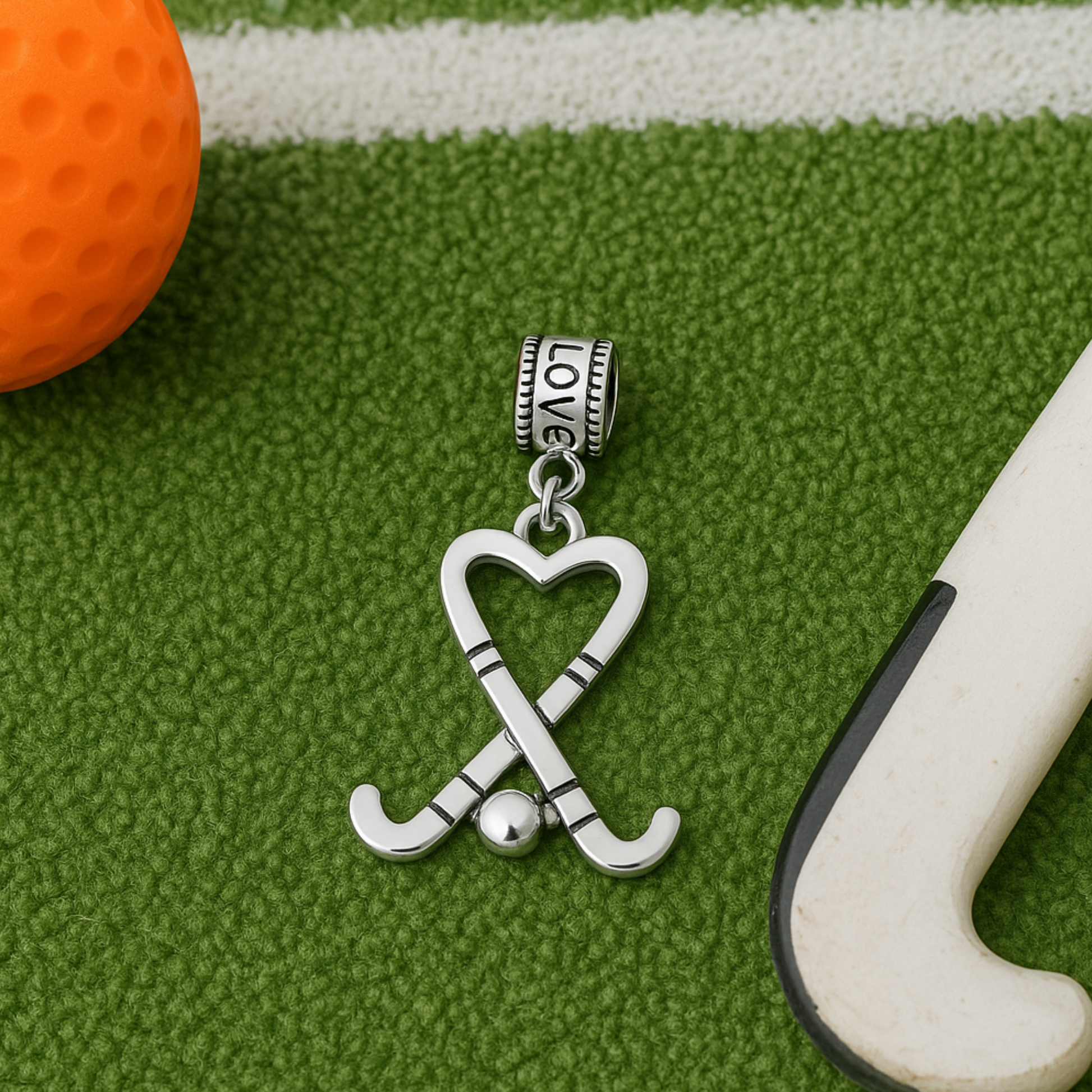 Silver crossed sticks field hockey charm bead placed on artificial turf with orange hockey bal