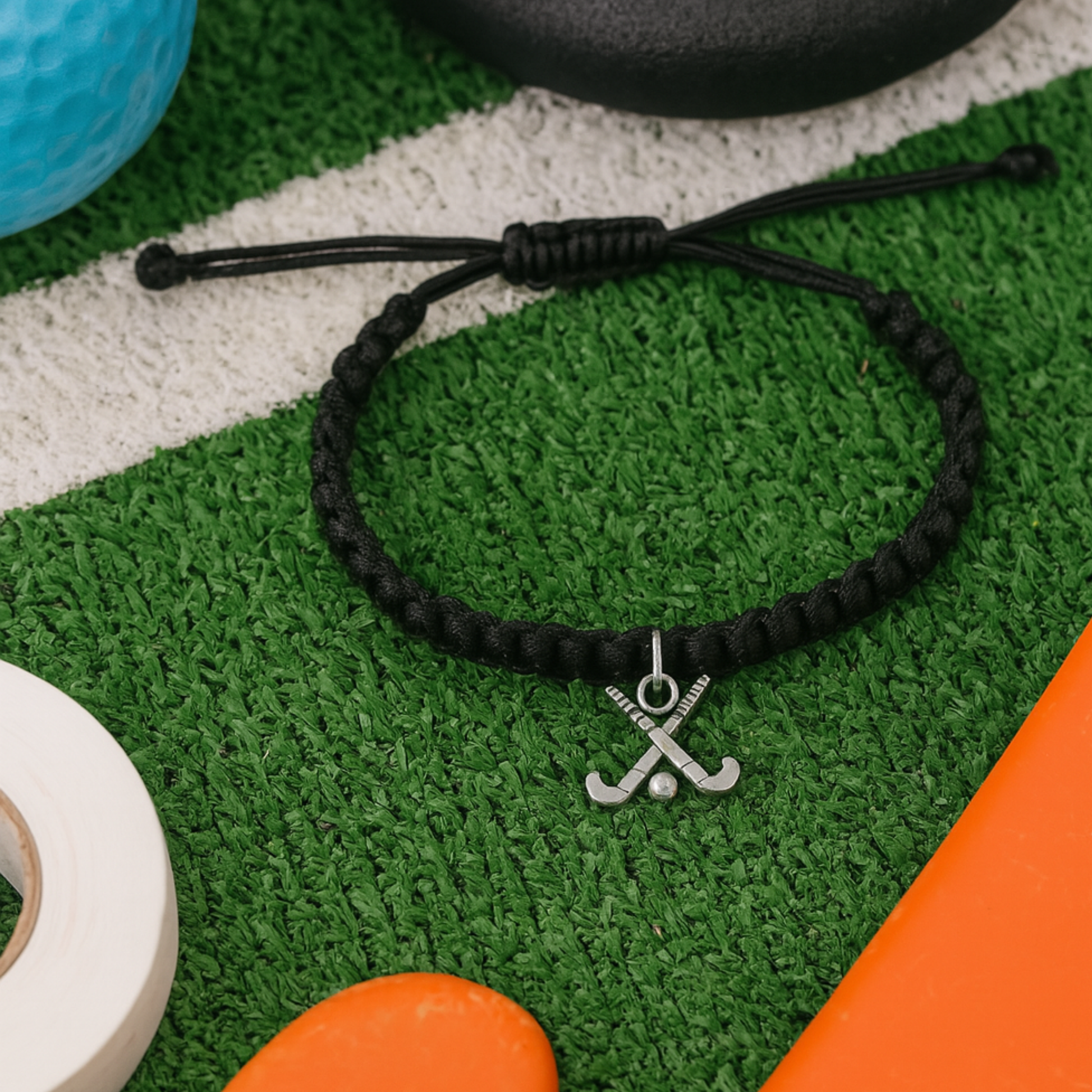 Black braided cord field hockey bracelet with silver charm displayed on turf with ball and stick
