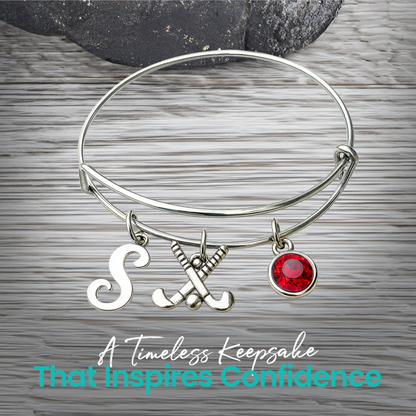 Silver adjustable field hockey bangle bracelet with personalized letter charm and red birthstone on rustic wood