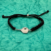 Black pickleball bracelet displayed on teal background, featuring silver-tone paddle charm and braided cord.

