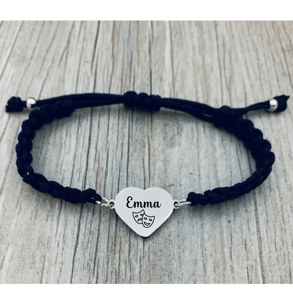 Lifestyle photo of black bracelet with engraved charm reading “Emma” and theater masks on wood background