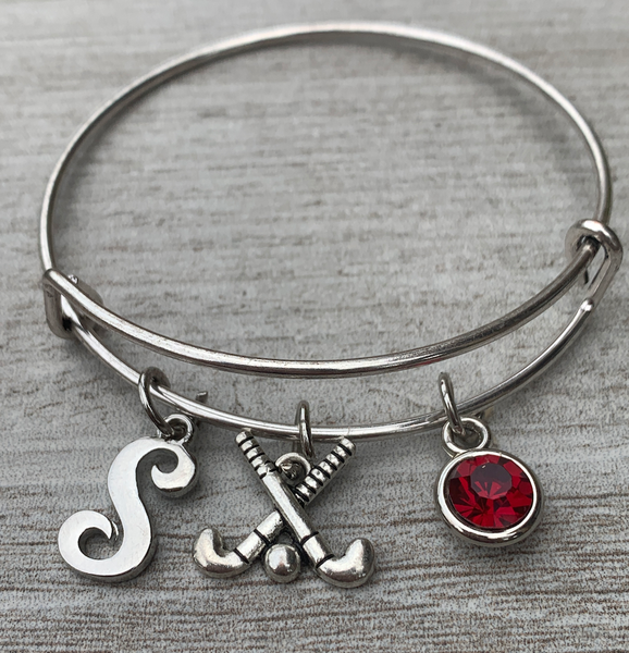 Personalized field hockey bangle bracelet with letter charm and red birthstone displayed on wooden background